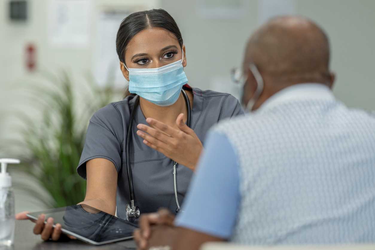 A medical professional wearing a face mask is explaining information on a computer tablet to an older man.