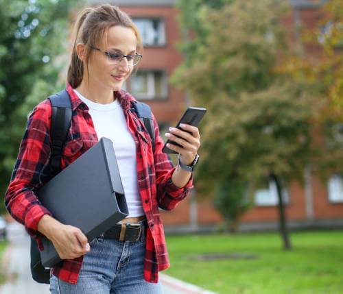 Female college student standing in front of a campus building looking at her smartphone.